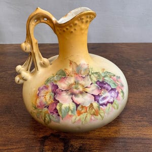 May include: A ceramic pitcher with a rounded body and a flared neck, featuring a gradient yellow to cream finish. The front is decorated with a painted floral arrangement of pink and purple blossoms with green leaves, and a gold-trimmed handle.