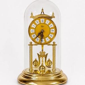 May include: A gold-tone Elgin anniversary clock encased in a clear glass dome. The clock features a yellow circular face with black Roman numerals, ornate black hands, and a rotating pendulum with three gold spheres supported by two pillars.