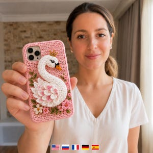 May include: A pink crochet phone case featuring a three-dimensional white swan with a small gold crown, accented by pink and white flowers and green leaves. The swan has a black eye and an orange beak, with layered white and pink wing feathers.