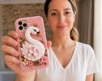 May include: A pink crochet phone case featuring a three-dimensional white swan with a small gold crown, accented by pink and white flowers and green leaves. The swan has a black eye and an orange beak, with layered white and pink wing feathers.