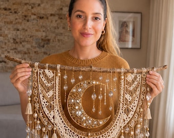 Può includere: Una decorazione da parete con base in ramo di legno naturale e corda in macramè color crema intrecciata in motivi complessi. Una mezzaluna dorata è decorata con cristalli e perline trasparenti, arricchita da gocce di cristallo pendenti.