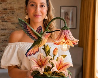 Può includere: Una lampada da tavolo all'uncinetto fatta a mano con un vivace colibrì dalle ali verdi, rosa e viola in volo, che si libra vicino a un grande fiore di giglio rosa rivolto verso il basso che funge da paralume, su una base con foglie verdi.