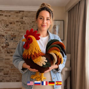 Puede incluir: Un gallo de peluche hecho a ganchillo con una cresta y barbillas de color rojo vibrante, cabeza amarilla y un cuerpo compuesto por plumas superpuestas en tonos marrón, verde oscuro y crema. El gallo tiene patas amarillas y cola marrón.