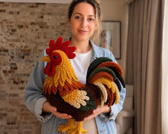 Può includere: Un gallo di peluche realizzato all'uncinetto con cresta e bargigli rosso vivo, testa gialla e un corpo composto da piume stratificate nei toni del marrone, verde scuro e crema. Il gallo ha zampe gialle e una coda marrone scuro.