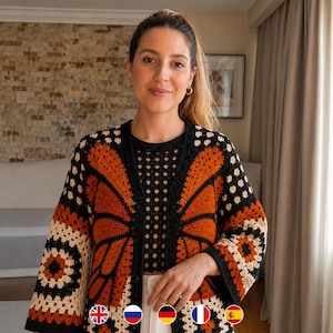 May include: A crochet cardigan featuring a butterfly wing pattern in burnt orange, black, and cream. The garment has long sleeves, an open front, and a square-patterned border. The design incorporates intricate stitch work throughout the fabric.