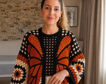 Può includere: Un cardigan all'uncinetto con un motivo ad ali di farfalla nei colori arancione bruciato, nero e crema. Il capo presenta maniche lunghe, apertura frontale e un bordo con motivo a quadrati. Il design include una lavorazione a maglia complessa.