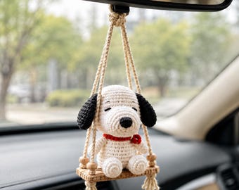 Può includere: Una figura di cane all'uncinetto bianca con orecchie nere e un collare rosso, seduta su una piccola altalena di legno sospesa da un cordino beige. L'ornamento presenta un piccolo dettaglio a fiore rosso sul collare per lo specchietto retrovisore.