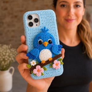 Könnte beinhalten: Eine hellblaue Handyhülle aus Häkelarbeit mit einem dreidimensionalen blauen Vogel mit weißem Bauch, orangefarbenem Schnabel und schwarzen Augen. Der Vogel sitzt auf einem braunen Ast, der mit kleinen rosa und weißen Blüten und grünen Blättern verziert ist.