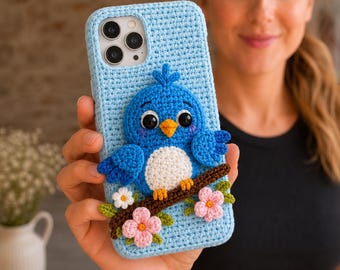 May include: A light blue crochet phone case featuring a three-dimensional blue bird with a white belly, orange beak, and black eyes. The bird sits on a brown branch decorated with small pink and white flowers and green leaves.