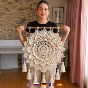 May include: A large, circular, off-white macrame wall hanging featuring an intricate, layered mandala pattern with detailed knotwork. The piece is suspended from a horizontal wooden dowel and finished with several hanging tassels at the bottom.