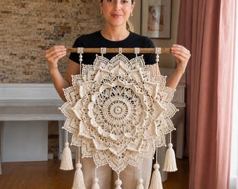 Può includere: Una grande decorazione da parete in macramè color bianco sporco, di forma circolare, caratterizzata da un intricato motivo a mandala a strati con dettagliati nodi. L'oggetto è appeso a un bastone di legno e rifinito con diverse nappe.