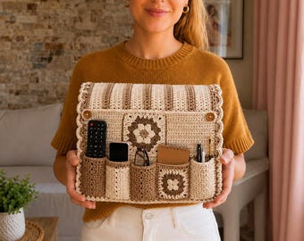 Può includere: Un organizer da parete rettangolare all'uncinetto con un motivo a righe beige e marrone e due decorazioni a granny square. Include cinque tasche verticali contenenti un telecomando, uno smartphone, occhiali, un taccuino e due penne.