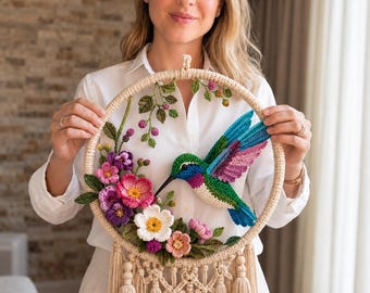 Può includere: Una decorazione da parete circolare con un colibrì all'uncinetto con ali verdi, blu e rosa, circondato da fiori all'uncinetto nei toni del rosa, viola e bianco, con foglie verdi, il tutto montato su un cerchio beige con nappe pendenti.