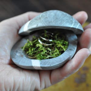 May include: A gray stone ring box with a white stripe, featuring a hinged lid that is partially open. The interior of the stone container is filled with green moss and holds two thin, metallic silver-colored rings resting on the moss.