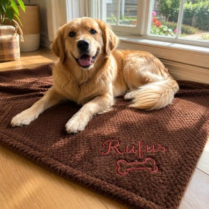 May include: A golden retriever dog lying on a dark brown textured blanket. The blanket features the embroidered name Rufus in red script lettering above a red embroidered dog bone shape. The dog has light golden fur and an open mouth expression.