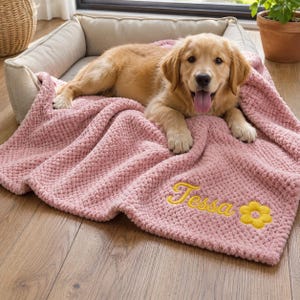 May include: A golden retriever puppy lying on a soft, textured, dusty pink blanket. The blanket features the embroidered name Tessa in yellow cursive script, accompanied by a small yellow flower icon with a brown center, stitched onto the fabric.