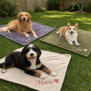 May include: Three square textured blankets in purple, olive green, and cream, each personalized with a dog's name and a small icon. The purple blanket features the name Toby with a fish bone, the green has Zola with a heart, and the cream has Moose with a heart.