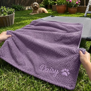 May include: A rectangular purple textured blanket featuring the embroidered name Daisy in a cursive script font, accompanied by a small embroidered paw print design located in the bottom right corner of the fabric.