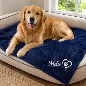 May include: A golden retriever dog resting on a navy blue textured blanket. The blanket features white embroidered text that reads Milo next to a white heart outline containing a paw print. The dog has light golden fur and an open mouth.