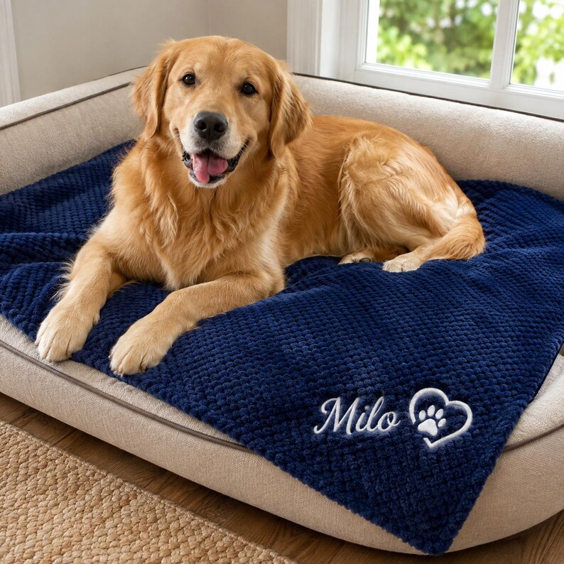 May include: A golden retriever dog resting on a navy blue textured blanket. The blanket features white embroidered text that reads Milo next to a white heart outline containing a paw print. The dog has light golden fur and an open mouth.