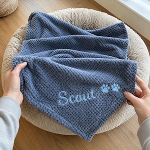 May include: A blue textured waffle-knit blanket featuring the embroidered name Scout in light blue script lettering, accompanied by two small embroidered light blue paw prints positioned to the right of the name on the fabric surface.
