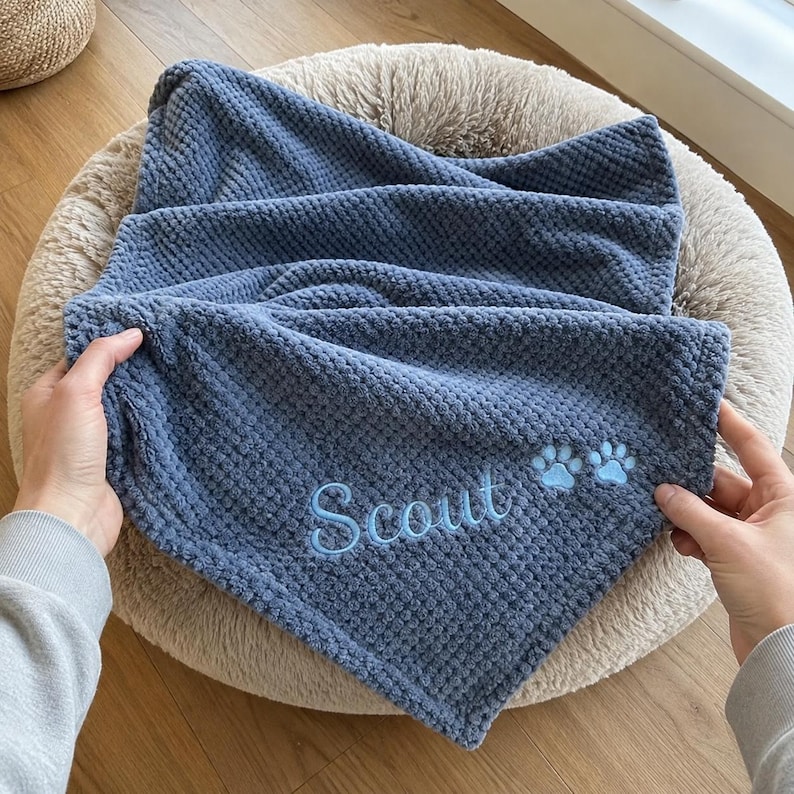 May include: A blue textured waffle-knit blanket featuring the embroidered name Scout in light blue script lettering, accompanied by two small embroidered light blue paw prints positioned to the right of the name on the fabric surface.
