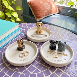 May include: Three round, speckled ceramic trinket dishes. One features a golden retriever figurine and the text SUNNY. Another shows a tortoiseshell cat figurine and the text OLIVE. The third displays a black pug and gray cat with the text BEAN MOCHI.