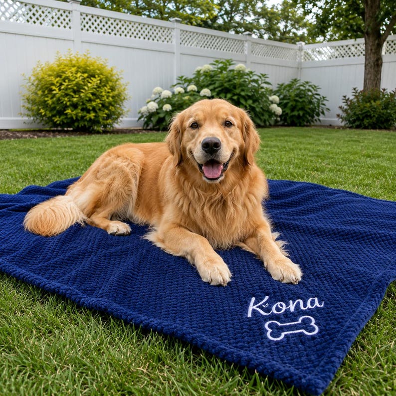May include: A golden retriever dog lying on a dark blue textured blanket. The blanket features white embroidered text that reads Kona above a white embroidered bone shape. The blanket has a waffle-like pattern and is spread out on green grass.