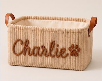 May include: A rectangular beige corduroy storage basket featuring the name Charlie in brown cursive lettering and a brown paw print icon. The basket has a cream-colored interior lining and two brown faux leather handles attached with gold rivets.
