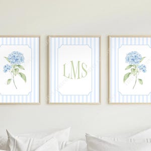 May include: A set of three framed wall art prints featuring a light blue vertical striped background. The two outer prints display a watercolor illustration of blue hydrangea flowers with green leaves. The center print features the letters LMS.