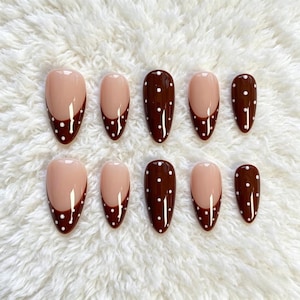 May include: A set of ten almond-shaped press-on nails featuring a glossy finish. The design combines a nude base with deep brown tips adorned with small white polka dots. The nails are arranged in two rows, showcasing varying sizes from thumb to pinky.