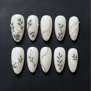 May include: A set of ten almond-shaped press-on nails with a glossy off-white base. The designs feature hand-painted green leafy vines, small white floral accents, delicate gold metallic lines, and tiny white pearl embellishments arranged in patterns.
