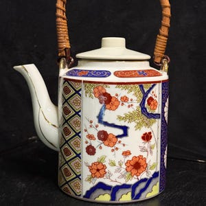 Puede incluir: Una tetera de porcelana blanca con cuerpo cilíndrico, asa de ratán trenzado y tapa. El exterior está decorado con coloridos motivos florales en naranja, rojo y azul, junto con patrones geométricos de diamantes y detalles en oro.