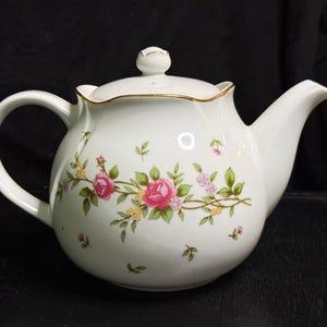 以下が含まれることがあります： ピンクのバラ、緑の葉、小さな黄色い花が描かれた繊細な花柄の白い磁器製ティーポット。ティーポットには金色の縁取りが施された波状の縁、湾曲したハンドル、つまみ付きの蓋、先細りの注ぎ口が付いています。
