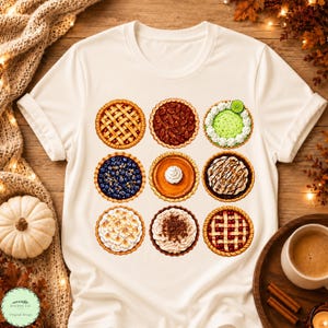 Puede incluir: Una camiseta de color crema con un estampado gráfico de nueve pasteles diferentes dispuestos en una cuadrícula. Los pasteles incluyen tartas de frutas con corteza de rejilla, una tarta de nueces, una tarta de lima con nata y una tarta de arándanos.