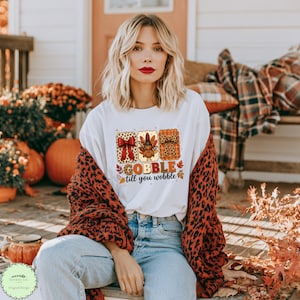 May include: A white short-sleeved t-shirt featuring a graphic print of three rectangular panels with a red bow, a turkey wearing a hat, and a leopard print bow. Below the panels, the text reads GOBBLE till you wobble in orange and brown.