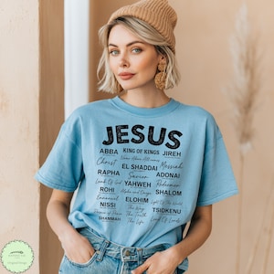 May include: A light blue short-sleeve t-shirt featuring black text that lists various names and titles for Jesus, including Abba, King of Kings, Jireh, Christ, El Shaddai, Messiah, Rapha, Savior, Adonai, Rohi, Yahweh, Redeemer, Emmanuel, Elohim, Shalom, Nissi, and more.