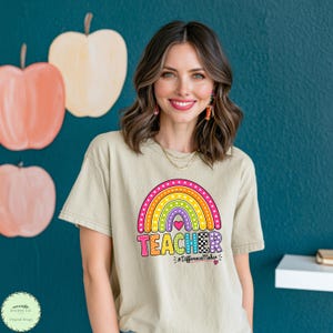 May include: A beige short-sleeve t-shirt featuring a colourful graphic of a rainbow with polka dots, stripes, and checkered patterns. Below the rainbow, the word TEACHER is printed in bold, multi-coloured letters, followed by #DifferenceMaker.