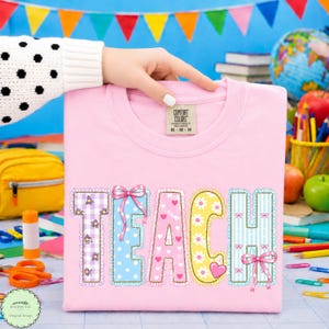Pode incluir: Uma t-shirt de gola redonda rosa claro com a palavra TEACH em letras grandes e decorativas. Cada letra tem um padrão único, incluindo xadrez vichy roxo com flores, bolinhas azuis com um laço rosa, corações rosa, margaridas amarelas e riscas azuis.