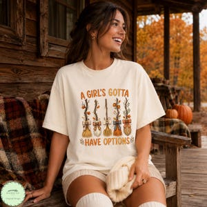 Può includere: Una t-shirt a maniche corte color crema con una grafica di cinque scope stilizzate decorate con foglie autunnali e fiocchi. Il testo "A GIRL'S GOTTA HAVE OPTIONS" è stampato in arancione sopra e sotto l'illustrazione delle scope.