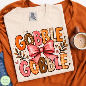 May include: A cream-colored t-shirt featuring the text GOBBLE GOBBLE in large, multi-patterned letters. The design includes a prominent pink bow in the center, surrounded by small hearts, star accents, and leaf illustrations in shades of orange, brown, and pink.