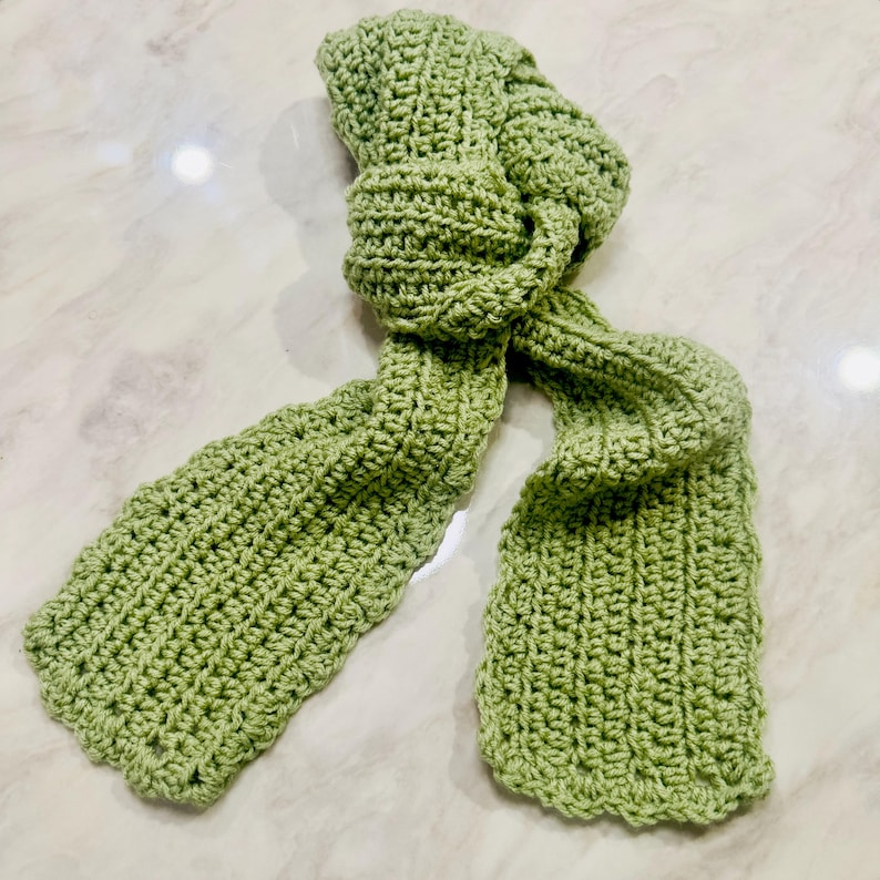 K&ouml;nnte beinhalten: Ein heller salbeigr&uuml;ner Schal mit einem strukturierten H&auml;kelstichmuster. Der Schal ist aus einer Reihe von miteinander verbundenen Schlaufen und Reihen gefertigt, wodurch ein konsistentes, sich wiederholendes, strick&auml;hnliches Aussehen entsteht.