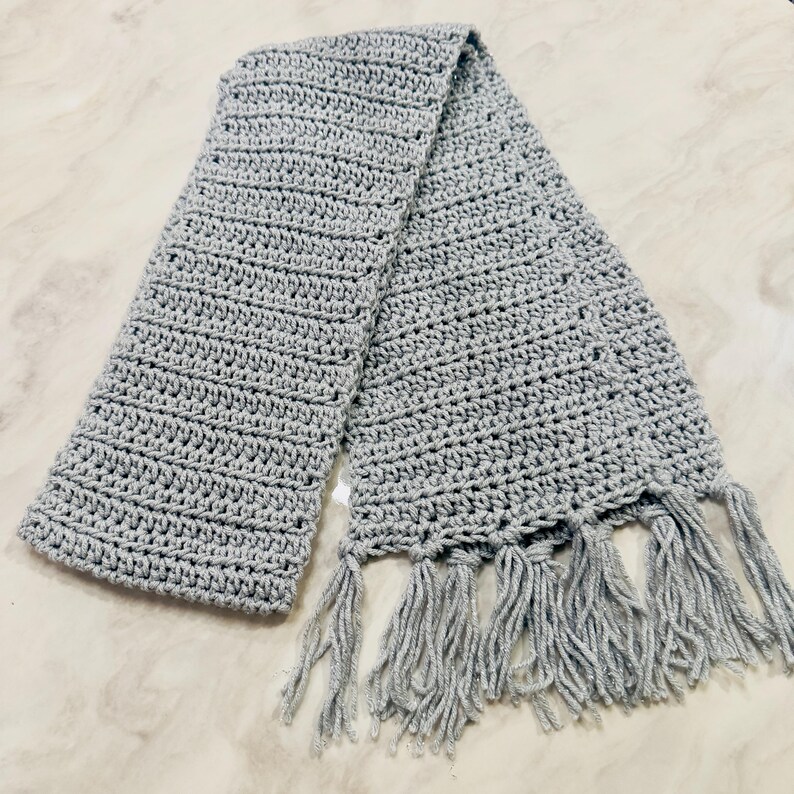 K&ouml;nnte beinhalten: Ein hellgrauer H&auml;kelschal mit einem durchgehenden, strukturierten Maschenmuster &uuml;ber die gesamte L&auml;nge des Stoffes. Der Schal ist mit einer Reihe langer, passender grauer Garnquasten versehen, die an der Unterkante befestigt sind.
