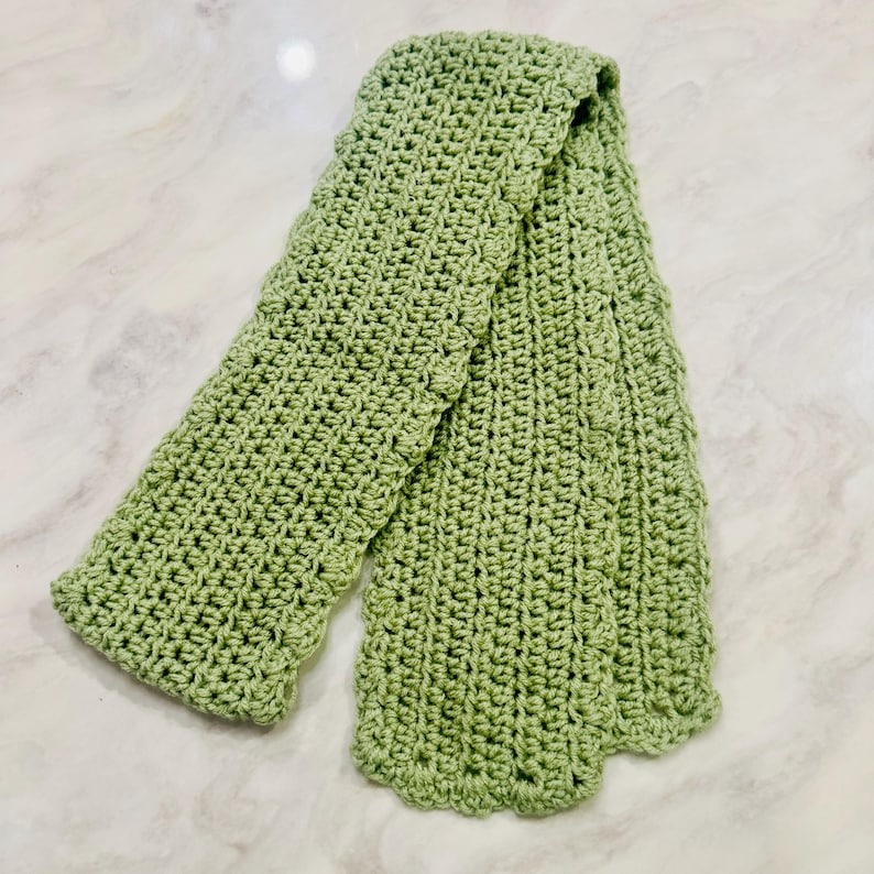 K&ouml;nnte beinhalten: Ein salbeigr&uuml;ner Schal mit einem strukturierten H&auml;kelstichmuster. Der Schal hat eine gleichm&auml;&szlig;ige rechteckige Form mit gewellten Kanten an den Seiten und Enden und weist eine einheitliche Strickkonstruktion &uuml;ber die gesamte L&auml;nge des Artikels auf.
