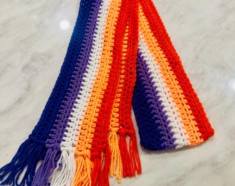Könnte beinhalten: Ein langer, rechteckiger Häkelschal mit einem lebendigen Farbverlauf aus dunkelblauem, violettem, weißem, orangefarbenem und hellrotem Garn. Der Schal ist in einem strukturierten Maschenmuster gefertigt und mit passenden Fransen an den Enden versehen.