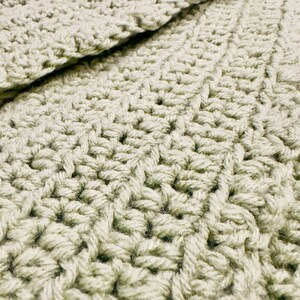 K&ouml;nnte beinhalten: Ein helles, salbeigr&uuml;nes geh&auml;keltes Textil mit einem dichten, sich wiederholenden Stichmuster. Der Stoff weist eine konsistente, strukturierte Oberfl&auml;che auf, die durch ineinandergreifende Garnschlaufen entsteht und eine einheitliche Farbe zeigt.
