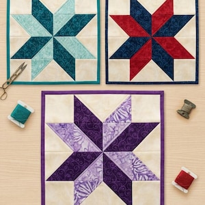 Peut inclure: Trois blocs de courtepointe carrés en tissu présentant des motifs d'étoiles à huit branches dans des tons turquoise, rouge et bleu marine, et violet. Chaque bloc a un fond crème, une bordure unie, des ciseaux et des bobines de fil.