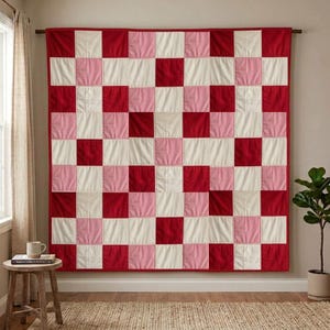 May include: A square patchwork quilt featuring a repeating grid pattern of alternating solid red, light pink, and off-white fabric squares. The quilt is hung flat against a wall, displaying a consistent checkerboard arrangement of these three colours.