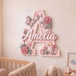 May include: A large, light pink wooden letter A wall decoration featuring the name Amelia in white script lettering. The letter is adorned with three-dimensional pink and white flowers, green leaves, and two butterflies in shades of pink and yellow.