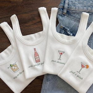 Puede incluir: Cuatro camisetas de tirantes blancas acanaladas con gráficos de cócteles bordados y texto. Los diseños incluyen una bebida con el texto easy on the ice, una botella de vino con corks are for quit, una copa de martini con pour decisions y un cóctel con just a splash.